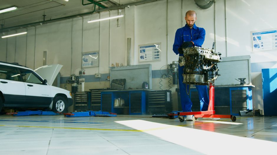 Mechanic lifting a car engine in a well-lit auto repair shop with vehicle in background.
